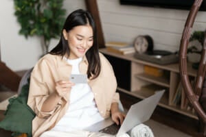 smiling korean girl buying online from home shopping laptop holding credit card