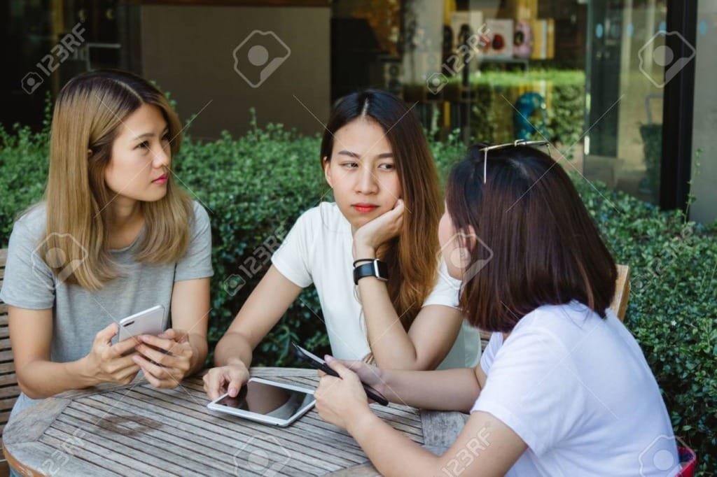 112963467 cheerful asian young women sitting in cafe drinking coffee with friends and talking together