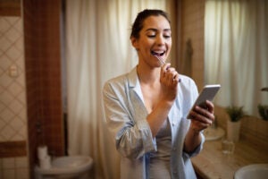 young woman texting mobile phone while brushing teeth bathroom